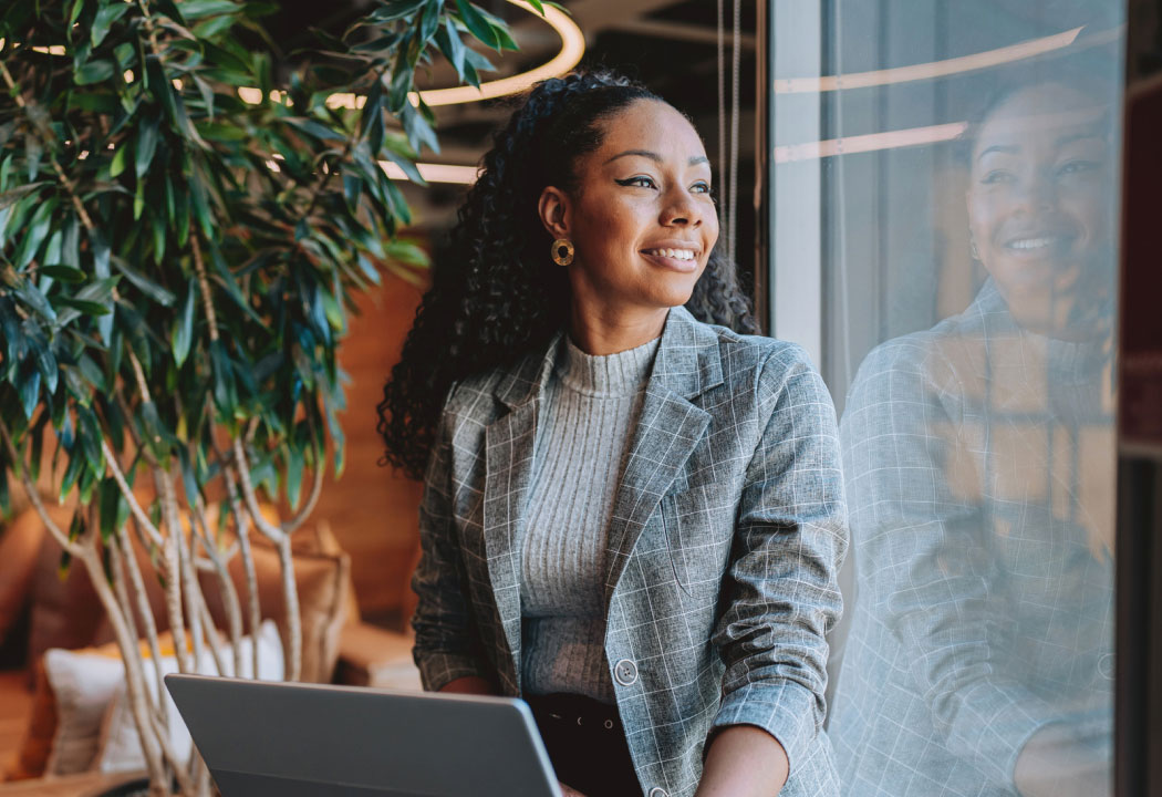 A young professional woman looking out a window and holding a laptop